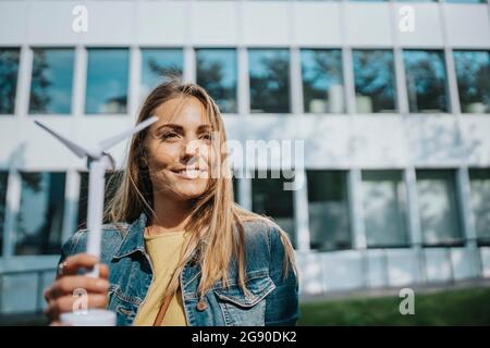 Frau in Denim-Jacke mit Windturbinenmodell vor dem modernen Bürogebäude Stockfoto