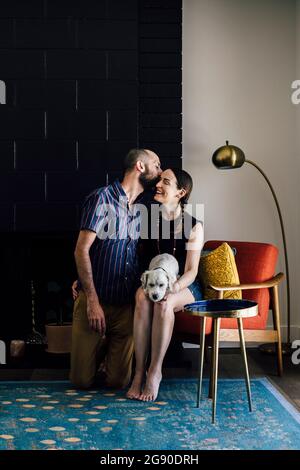 Fürsorglicher Mann küsst Frau mit Hund sitzt auf einem Stuhl im Wohnzimmer Stockfoto