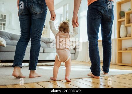 Baby Mädchen hält die Hände der Eltern, während sie im Wohnzimmer spazieren Stockfoto