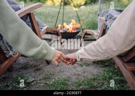 Junges Paar, das sich die Hände hält, während es auf einem Stuhl in der Nähe der Feuerstelle sitzt Stockfoto