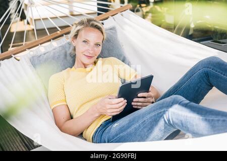 Frau mit digitalem Tablet lächelt, während sie sich in der Hängematte auf dem Balkon entspannt Stockfoto