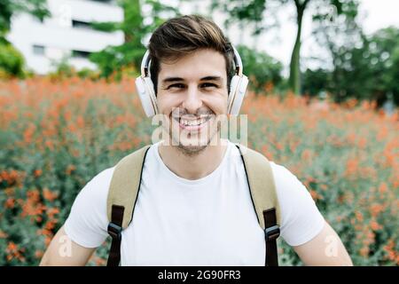 Lächelnder junger Mann, der über Kopfhörer Musik hört, mitten auf der Wiese Stockfoto