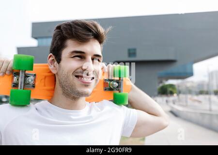 Lächelnder junger Mann, der Skateboard auf der Schulter trägt, während er wegschaut Stockfoto