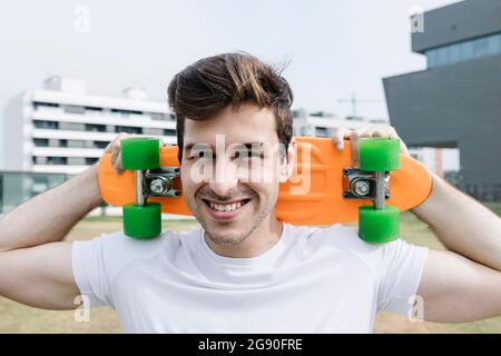 Lächelnder junger Mann, der Skateboard auf der Schulter trägt Stockfoto