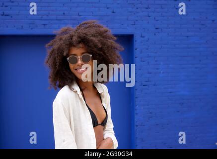 Lächelnde junge Afro-Frau mit Sonnenbrille vor der blauen Wand Stockfoto