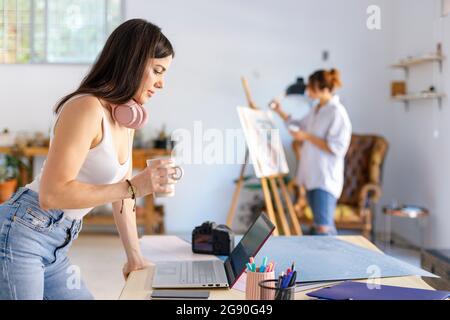 Fotografin hält den Becher auf dem Schreibtisch, während die Künstlerin im Hintergrund malt Stockfoto