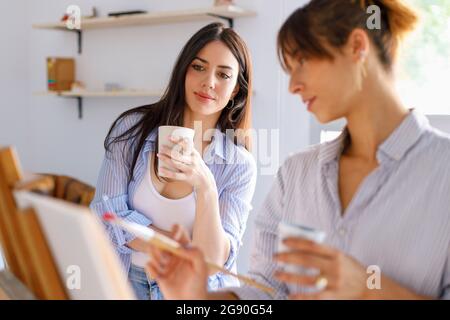 Künstlerin mit Tasse und Blick auf die Malerei von Kollegen im Atelier Stockfoto