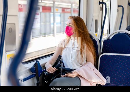 Geschäftsfrau in schützender Gesichtsmaske, die durch das Fenster schaut, während sie im Zug pendelt Stockfoto