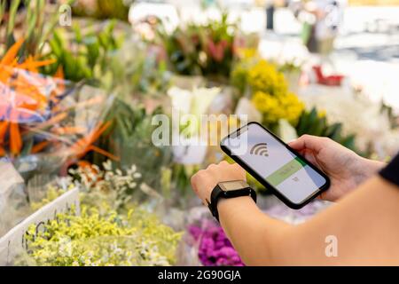 Frau, die kontaktloses Bezahlen über Smartwatch und Mobiltelefon im Blumenladen macht Stockfoto