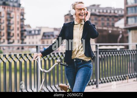 Fröhliche Frauen, die professionell Musik über Kopfhörer hören, während sie mit dem Fahrrad auf der Brücke fahren Stockfoto