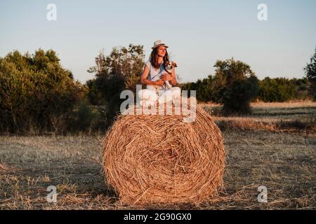 Frau, die während des Sonnenuntergangs mit Hund auf Strohballen sitzt Stockfoto