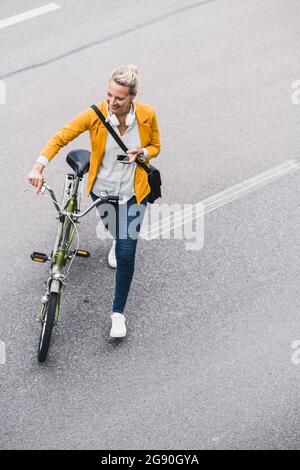 Glückliche weibliche Pendlerin, die das Mobiltelefon hält, während sie auf der Straße mit dem Fahrrad fährt Stockfoto