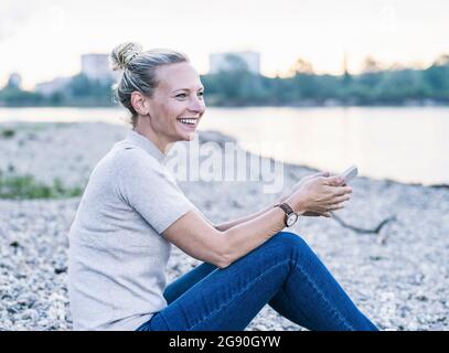 Glückliche, reife Frau, die am Flussufer mit dem Mobiltelefon sitzt Stockfoto