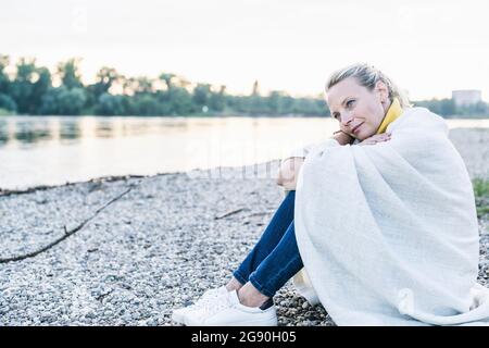 Nachdenkliche Frau, die in eine Decke gehüllt ist und auf Kieselsteinen am Flussufer sitzt Stockfoto