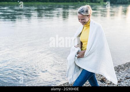 Lächelnde Frau mit Decke, die am Flussufer entlang läuft Stockfoto