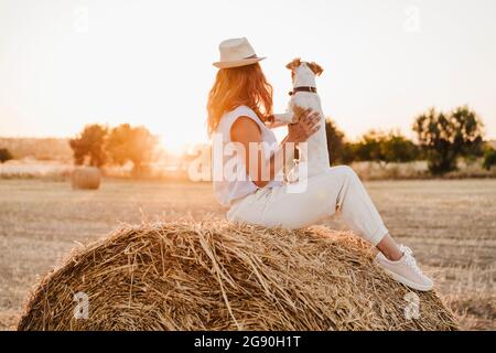 Frau mit Hund sitzt auf Strohballen auf der Farm während des Sonnenuntergangs Stockfoto