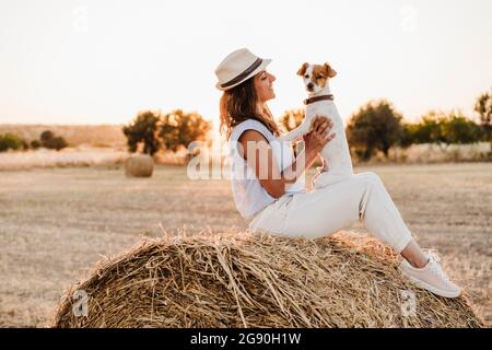 Lächelnde Frau, die beim Sonnenuntergang auf Strohballen sitzt und den Hund ansieht Stockfoto