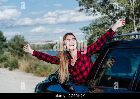 Fröhliche junge Frau mit ausgestreckten Armen, die sich vom Autofenster lehnt und den sonnigen Tag genießen Stockfoto