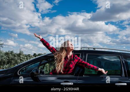 Glückliche blonde Frau mit ausgestreckten Armen, die sich an sonnigen Tagen vom Autofenster lehnt Stockfoto