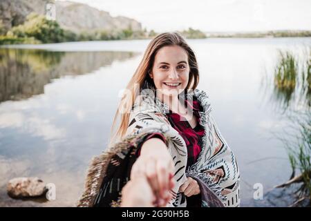 Glückliche junge Frau, die in eine Decke gewickelt war und die Hand einer Freundin am Seeufer hielt Stockfoto