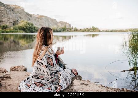 Junge blonde Frau in eine Decke gewickelt, die Kaffeebecher am Seeufer hält Stockfoto