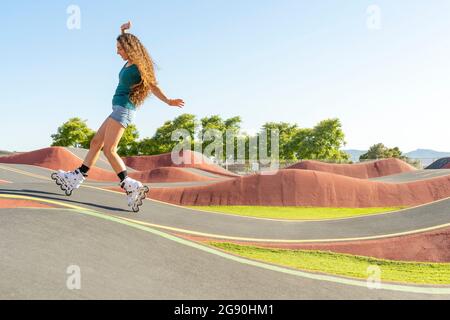 Junge Frau mit braunen Haaren genießt Inline-Skating auf der Pumptrack an sonnigen Tag Stockfoto