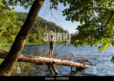 Mann, der an sonnigen Tagen auf einem umgestürzten Baum am Seeufer steht Stockfoto