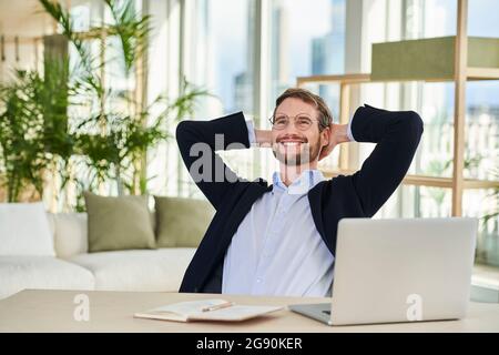 Fröhlicher entspannter männlicher Freiberufler, der im Heimbüro mit den Händen hinter dem Kopf sitzt Stockfoto
