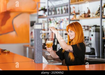 Lächelnde Rothaarige Kellnerin, die an der Bar ein Bierglas vom Fass füllte Stockfoto