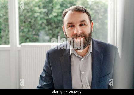 Lächelnder bärtiger, reifer Geschäftsmann, der im Büro sitzt Stockfoto