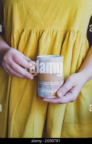 Eine junge, weiße Frau in einem einfachen, gelben Leinenkleid hält in ihren Händen eine Tonerde-Tasse aus Keramik in einer originellen, handgefertigten Form. Speicherplatz kopieren. Trend. Stockfoto