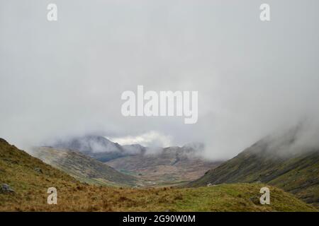 Ein nebeliges Bergtal: Wolken hängen tief um Berggipfel; Gras im Vordergrund; ein Tal mit einer Straße, die sich hinabschlängelt - Kurven auf der Route quer Stockfoto