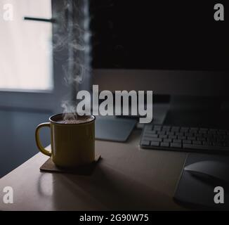Eine gelbe Tasse Kaffee steht auf dem Schreibtisch im sonnigen Morgen-Abend-Licht, neben dem Monitor und der Tastatur. Stockfoto
