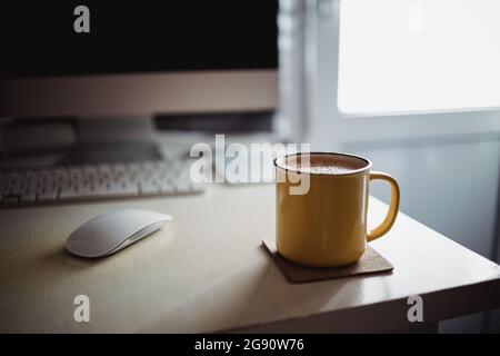 Eine gelbe Tasse Kaffee steht auf dem Schreibtisch im sonnigen Morgen-Abend-Licht, neben dem Monitor und der Tastatur. Stockfoto