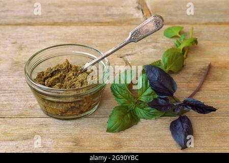 Basilikum-Pesto in Glasschale mit Metalllöffel und grünen lila Blättern auf Holztisch Hintergrund oben Stockfoto