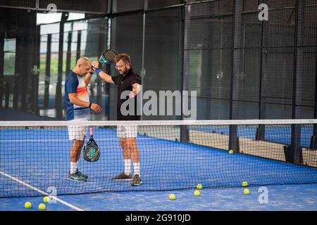 Überwachen Sie den Padel-Unterricht für den Mann, sein Schüler - Trainer lehrt Jungen, wie Padel auf dem Hallentennisplatz zu spielen Stockfoto