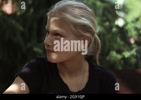 Ein blondes Teenager-Mädchen in einem schwarzen T-Shirt sitzt im Grünen und schaut mit Interesse davon. Porträt eines hübschen blonden Mädchens im Sonnenlicht. Stockfoto