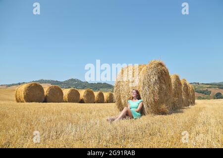 Das italienische Mädchen sitzt mit dem Rücken gegen einen Heuhaufen und genießt die Wärme der Sonne auf einem Bauernhof Stockfoto