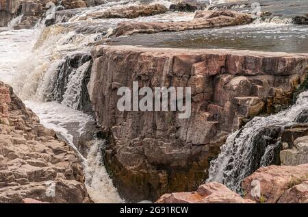 Sioux Falls, SD, USA - 2. Juni 2008: Nahaufnahme von braun-schwarzem Gestein, der von einem Wasserüberlauf verschont wird, durch das spritzende weiße Wasser fließt. Stockfoto