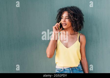 Horizontales Porträt einer afro-Frau, die mit ihrem gelben Smartphone anruft und lächelt. Sie lehnt sich an eine grüne Wand und verwendet Sommerkleidung Stockfoto