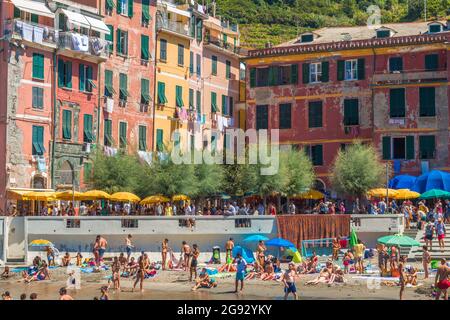 Touristen und Einheimische sonnen sich am kleinen Strand mit bunten Gebäuden im Hintergrund, aufgenommen in Vernazza, Cinque Terre, Italien. Stockfoto