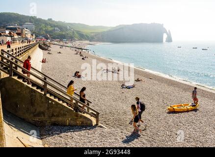 Touristen genießen den Kiesstrand in Etretat, Normandie, mit der Aval-Klippe, seinem Bogen und der Nadel in der Ferne. Stockfoto