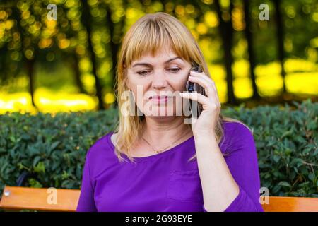 Unschärfe-Eleganz kaukasische blonde Frau reden, sprechen auf dem Telefon draußen, im Freien. 45er Jahre alte Frau in lila Bluse im Park. Erwachsene Frauen Stockfoto