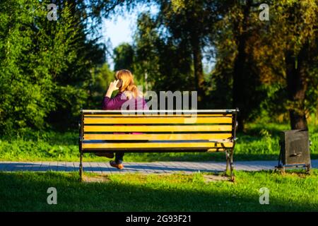 Unschärfe kaukasisch blonde Frau reden, sprechen am Telefon draußen, im Freien. 45er Jahre alte Frau in lila Bluse im Park auf der Bank. Erwachsene Frauen Stockfoto