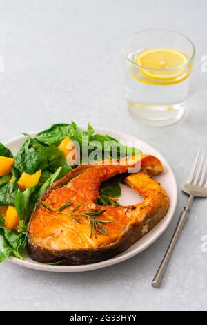 Gebratenes Lachssteak mit Spinat und gelben Tomaten auf einem Teller auf grauem Grund, ein Glas Wasser mit Zitrone, ein herzhaftes Mittagessen mit gesundem rotem Fisch Stockfoto