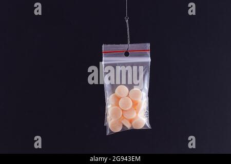 Die pharmazeutische Falle und Drogensucht - farbige Pillen oder Tabletten, die in einem kleinen Ziplock-Beutel auf einem Angelhaken gegen den schwarzen Hintergrund hängen Stockfoto