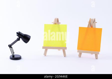 Zwei hölzerne Easels Miniature mit blanker quadratischer Leinwand oder Memopapier - Mockup. Mini-Holzständer mit sauberer Tafel und kleiner schwarzer Tischleuchte auf weißem Hintergrund, Kopierraum Stockfoto