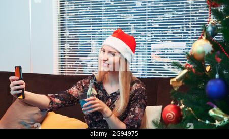 Seitenansicht der gut aussehenden Frau, die selfie in Santa Hut und mit Getränk in den Händen macht. Im Hintergrund stehen ein Weihnachtsbaum und Wolkenkratzer. Stockfoto