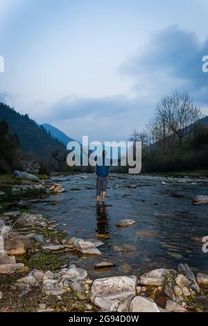 Junger Mann, der im Wasserbach mit Bergtal und Waldblick am Morgen aus flachem Winkel steht, wird am Shergaon arunachal pradesh india aufgenommen Stockfoto
