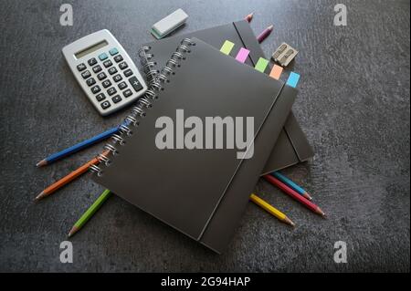 Schwarze Spiralhefte, bunte Buntstifte, Taschenrechner und stationäres Zubehör auf einem dunkelgrauen Schreibtisch, Kopierraum, ausgewählter Fokus, enge Schärfentiefe Stockfoto